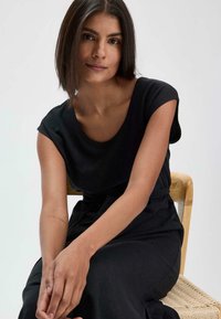 Musta mekko lyhyillä hihoilla ja pyöreällä kaula-aukolla, jossa on kerätty vyötärö. Sileä kangas rentoa istuvuuden kanssa, yhdistettynä puutuoliin.