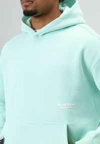 Mintgroene hoodie met een voorzak, zachte textuur en subtiele witte branding. Heeft een capuchon met trekkoord en geribbelde boorden.