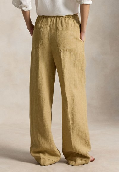 Άτομο που φοράει χαλαρό, μπεζ παντελόνι λινό με ελαστική μέση και πίσω τσέπες, στέκεται με τα χέρια στις τσέπες μπροστά σε ουδέτερο φόντο.