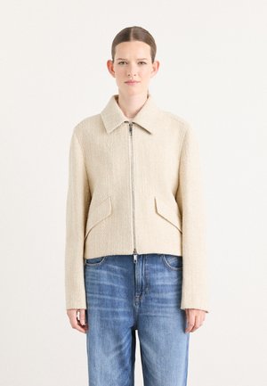 Giacca beige con zip, in tessuto testurizzato, colletto aperto, due tasche frontali e design corto, abbinata a jeans azzurri chiari.