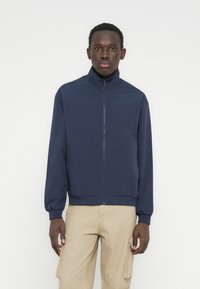 Άνδρας που φοράει μπλε ναυτικό σακάκι με φερμουάρ και μπεζ παντελόνι cargo, στέκεται μπροστά σε απλό λευκό φόντο.