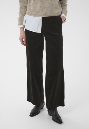 Persona que lleva pantalones de pana negros de pierna ancha, un suéter beige sobre una camisa blanca y botines negros de tacón sobre un fondo blanco.