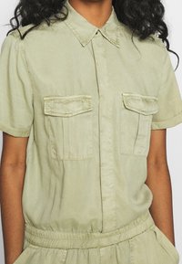 Ljusgrön kortärmad skjorta med spetsig krage, två bröstfickor och rynkad midja, med en slät tygtextur.