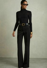 Top negro de cuello alto combinado con pantalones negros de cintura alta. Acabado con un cinturón ancho negro con hebilla dorada. Gafas de sol de ojo de gato.