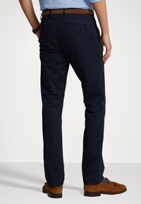 Pantaloni blu navy realizzati in un tessuto liscio, con un classico taglio dritto e tasche posteriori con chiusura a bottone. Cintura e scarpe marroni.