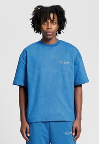 Sininen ylipitkä t-paita, jossa on pyöreä kaula-aukko, lyhyet hihat ja teksturoitu pinta. Vasemmassa rinnassa pieni valkoinen brodeerattu logo.