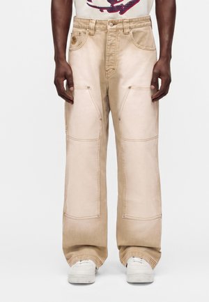 Pantalon beige clair à jambes larges avec renforts aux genoux, porté avec des baskets blanches, présenté de la taille aux pieds sur une personne à la peau foncée.