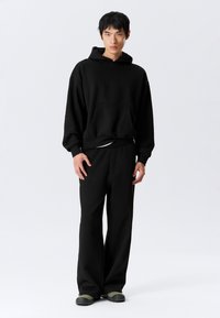 Black hooded sweatshirt with front pocket, paired with loose-fitting black pants. Fabric appears textured and soft, with relaxed silhouette.