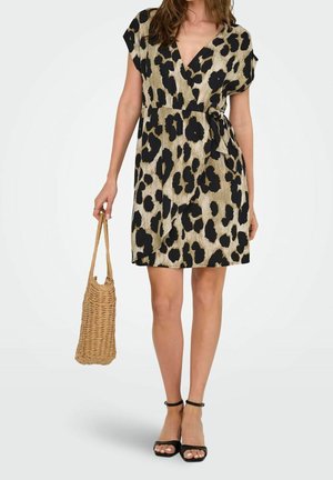 Femme portant une robe portefeuille courte à imprimé léopard beige et noir, tenant un sac en paille tressée, et chaussant des sandales noires à bride autour de la cheville.