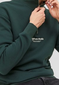Osoba v tmavozelené mikině s částečným zipem, ruce upravující zip, viditelný bílý nápis "Originals Studio" na hrudi.
