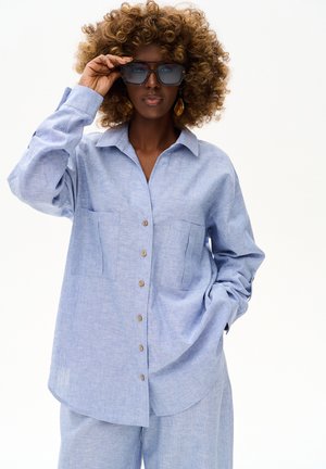 Kobieta z kręconymi włosami, ubrana w oversize niebieską koszulę na guziki i dopasowane spodnie, poprawia jedną ręką duże kwadratowe okulary przeciwsłoneczne.
