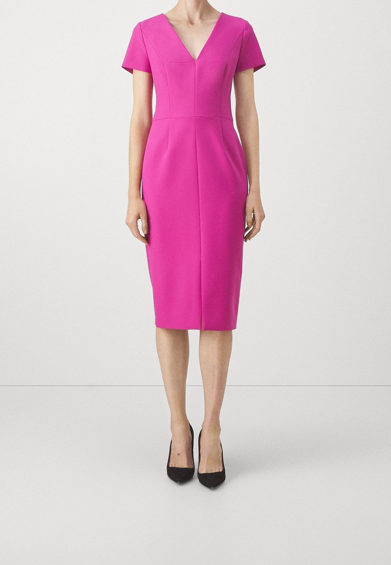 Femme portant une robe ajustée rose vif longueur genoux avec des manches courtes et des escarpins noirs à bouts pointus, debout devant un fond blanc uni.