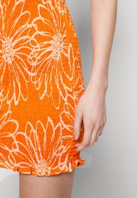 Oranžové květinové šaty s texturovanou, nařasenou látkou a bílými květinovými vzory. Rukou opřenou o bok šatů.