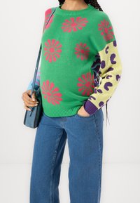 Pull vert en tricot avec des motifs floraux roses, manches contrastantes en jaune et violet, associé à un jean bleu à jambes larges.