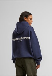 Mørkeblå oversized hettegenser med snor. Har "FORBUDT" med fet hvit skrift på ryggen med liten tekst under.