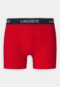 Sarkanas kokvilnas bokseršortus ar melnu jostu, uz kuras ir baltā "Lacoste" zīmola apdruka. Gluda virsma, pieguļošs dizains un garums līdz vidus augšstilbam.
