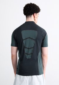 Camisa desportiva preta e verde-escura com painéis traseiros texturizados em contraste, mangas curtas e decote em crew, projetada para vestuário de performance.