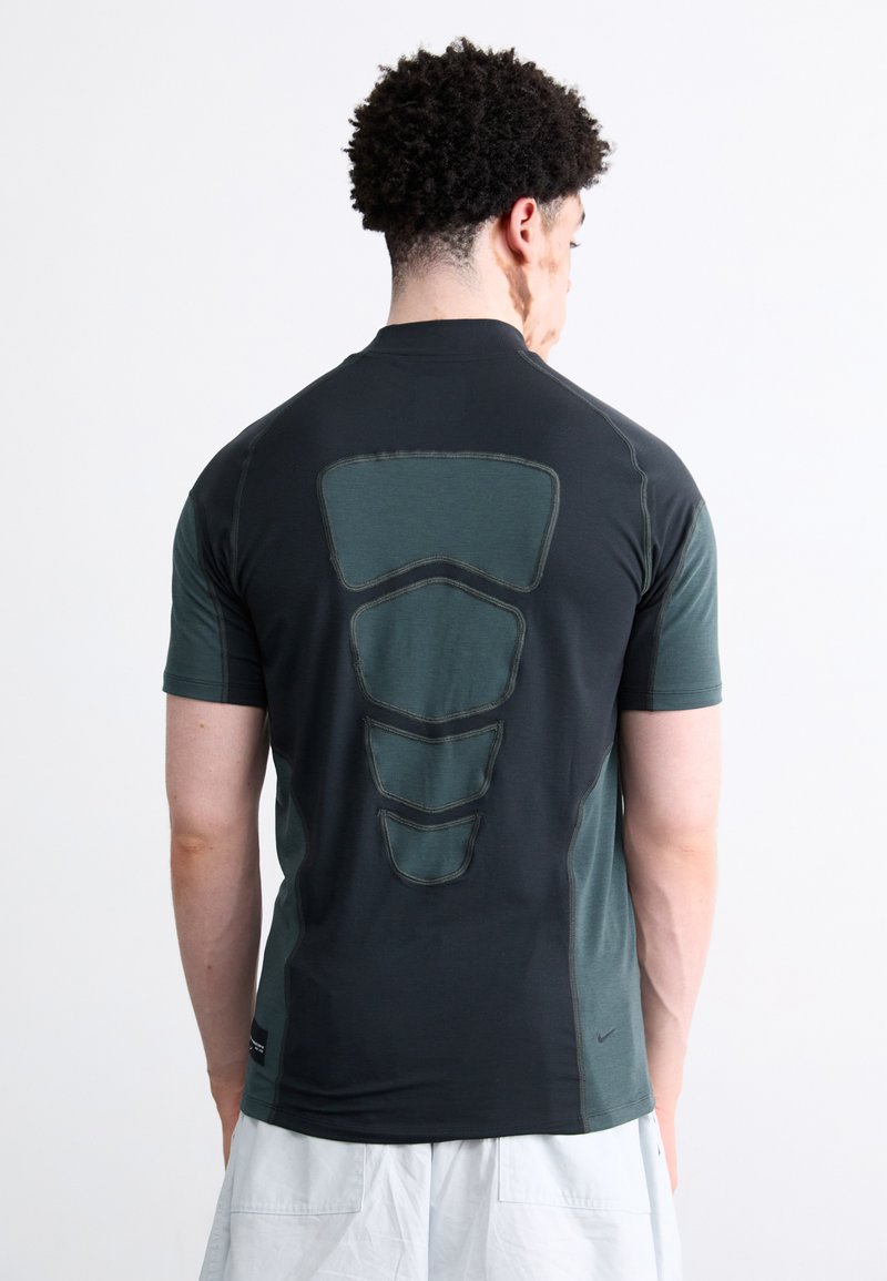 Camisa desportiva preta e verde-escura com painéis traseiros texturizados em contraste, mangas curtas e decote em crew, projetada para vestuário de performance.