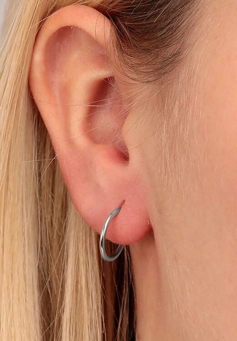 Silberne Creolen, kreisförmiges Design, dünnes Profil, polierte Oberfläche, getragen in einem durchstochenen Ohrläppchen, mit natürlicher Haarstruktur im Hintergrund.
