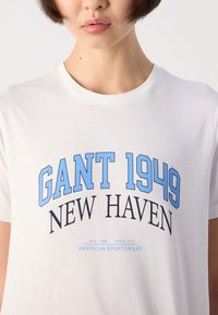 Asmuo vilkintis baltą marškinėlį su mėlynu ir juodu tekstu, rašančiu "GANT 1949 NEW HAVEN Amerikos sportinė apranga" ant krūtinės.