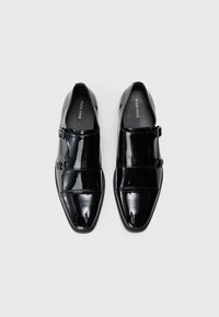 Schwarze Lacklederschuhe mit einer spitzen Zehenpartie, zwei Riemenschnallen und einer glatten, glänzenden Oberfläche. Flache Sohle, minimalistisches Design.