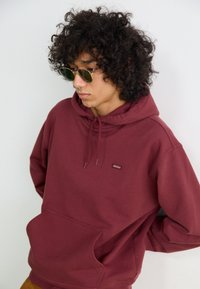 Maroon hoodie with a front pocket, drawstring hood, and a small logo patch. Model wears round sunglasses, has curly hair.