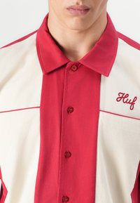 Rød og kremfarget kortermet skjorte med knapper og krage, med en brystlomme og brodert "Huf"-logo i rødt. Glatt stofftekstur.
