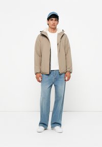 Beige puffjakke med faux fur-foring, lynlåsåbning foran, båret over en hvid t-shirt, kombineret med lyseblå brede bukser og hvide sneakers.