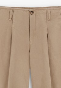 Pantalones beiges de corte amplio, hechos de algodón suave. Cuentan con cierre de botón y dos bolsillos frontales, mostrando costuras cosidas y un acabado texturizado.