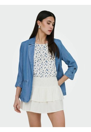 Femme aux longs cheveux bruns portant un blazer bleu, un chemisier blanc à motifs floraux et une jupe blanche à volants, posant devant un fond clair uni.