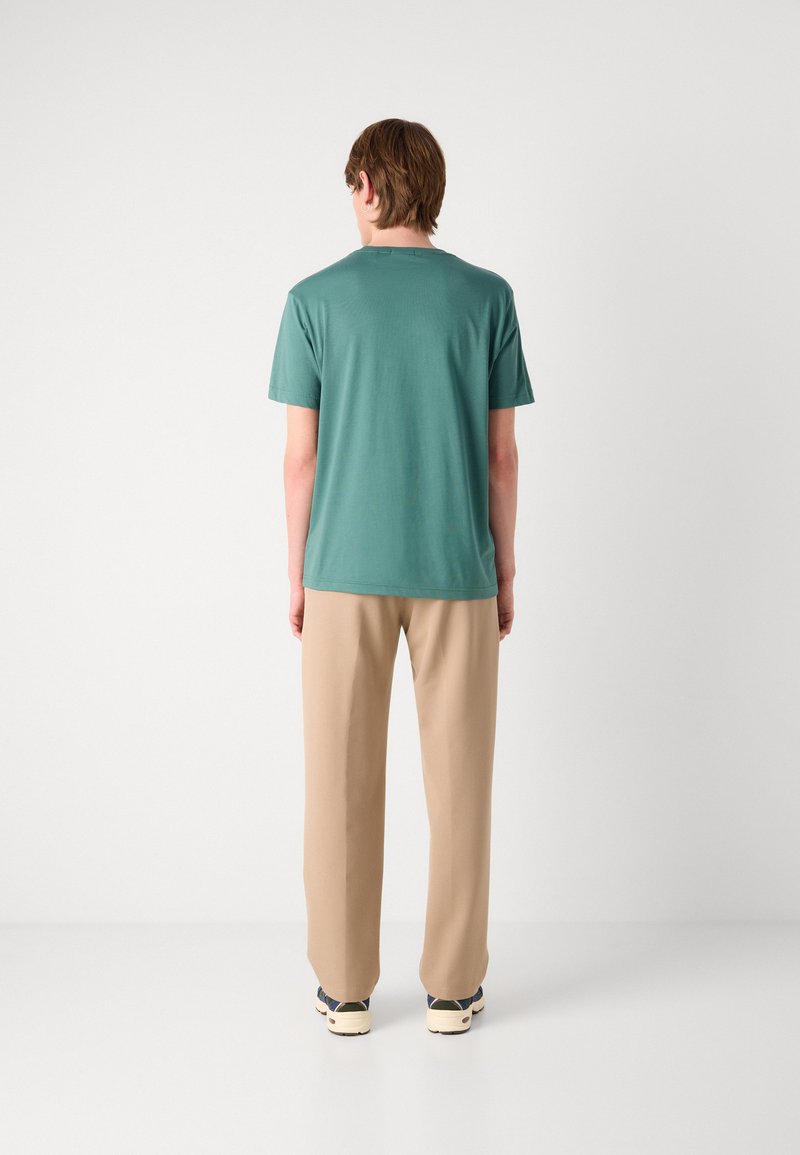 Türkise kurzärmelige T-Shirt mit Rundhalsausschnitt, kombiniert mit hellen beigen Hosen, auf einem schlichten Hintergrund stehend. Lässige Passform und glatte Textur.