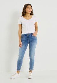 Vaqueros ajustados azules con un lavado descolorido, con cierre frontal de botón y trabillas para cinturón. Combinados con una camiseta blanca de manga corta con escote en V y zapatillas blancas.