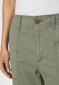 Olivgröna byxor med knäppning, prydda med framfickor, synlig sömnad och en jämn yta. Närbild som framhäver designens detaljer.