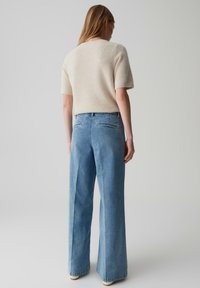 Vrouw die wegkijkt, gekleed in een crèmekleurige gebreide trui met korte mouwen en hoge taille wijdvallende blauwe jeans, tegen een lichtgrijze achtergrond.