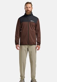 Giacca zip-up marrone e nera con cappuccio, caratterizzata da un logo sul petto, abbinata a pantaloni beige chiaro e scarpe da trekking grigie.