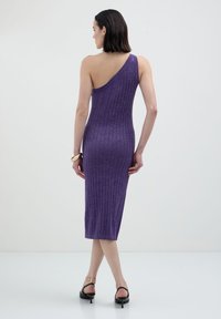 Donna in un vestito midi viola a coste con una spalla sola, con tacchi neri, bracciale alla caviglia dorato e bracciale dorato al polso, in piedi contro uno sfondo semplice.