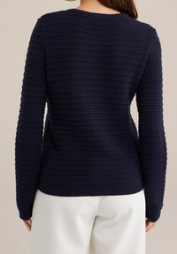 Granatowy sweter dziany z teksturowanym, paskowanym wzorem, długimi rękawami oraz prążkowanym dekoltem i dołem. Noszony na jasnych spodniach.
