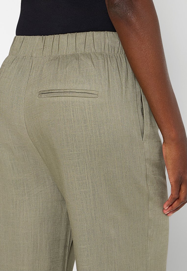 Nahaufnahme einer Person, die helle olivfarbene elastische Hose mit Schlitz-Rückentasche und seitlicher Nahttasche trägt, gekleidet in einem schwarzen Oberteil.