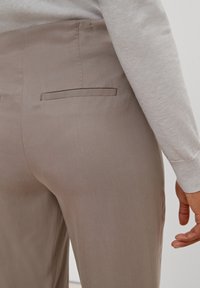 Pantalons ajustés marron clair vus de l'arrière, avec une poche arrière unique et un tissu lisse et texturé. Un pull gris décontracté est visible.