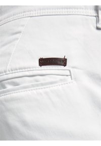 Bílá textilie s hladkým povrchem a prošitými detaily, s hnědou koženou nášivkou "JACK & JONES" připevněnou poblíž kapsy.