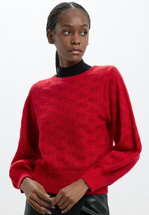 Maglione rosso lavorato a maglia con motivo testurizzato e maniche ampie, dotato di un collo a coste nero. Il materiale sembra morbido con una leggera lucentezza.