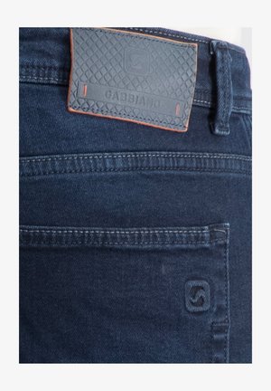 Tmavě modré džíny z denimu s prošitým zadním kapsou a obdélníkovou koženou značkovou nášivkou s texturovaným vzorem na pásku.