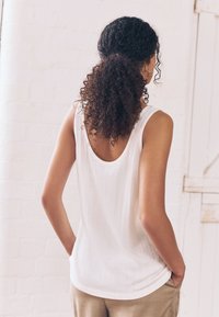 Hvid tanktop med et tekstureret ribbet mønster, rund halsudskæring og en let scoop på bagsiden, parret med beige bukser.