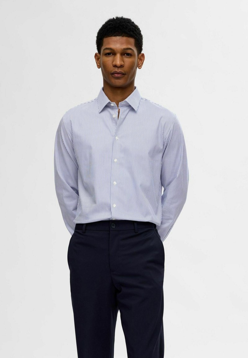 Camicia a righe blu e bianche con bottoni, in cotone, con colletto a punta; abbinata a pantaloni eleganti scuri.