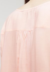 Blusa rosa chiaro realizzata in tessuto morbido e fluido. Presenta dettagli di pieghe sul retro e un collo rotondo, creando una silhouette rilassata.