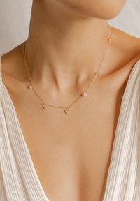 Collier en chaîne dorée avec cinq accents en forme de fleur, chacun orné d'un petit bijou. Le design est délicat et s'adapte à un décolleté mis en valeur par le tissu.