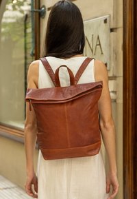 Brązowy skórzany plecak z regulowanymi paskami, składanym klapem i zamknięciem na zamek. Gładka tekstura z minimalnymi detalami w szyciu.