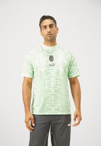 Camiseta de manga corta verde claro con un patrón geométrico por toda la prenda, con cuello redondo y acentos de logo negro en el pecho.