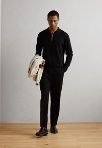 Suéter negro con cuello de cierre, pantalones negros y zapatos negros. El modelo sostiene una chaqueta beige. Tela suave con un corte ajustado, de pie sobre un suelo de madera.