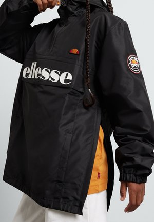 Personne portant une veste noire ellesse avec un logo sur la poitrine et la manche, dézippant partiellement le col montant, montrant un vêtement orange en dessous.